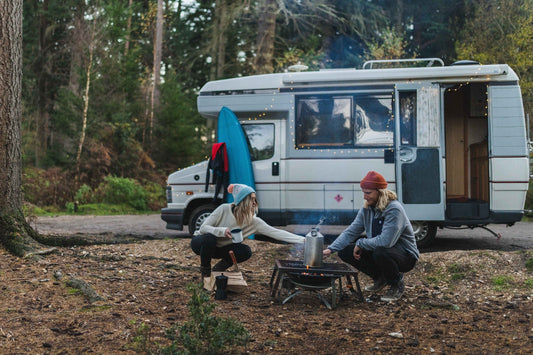 A man and a woman cook on a campfire at the side of their campervan